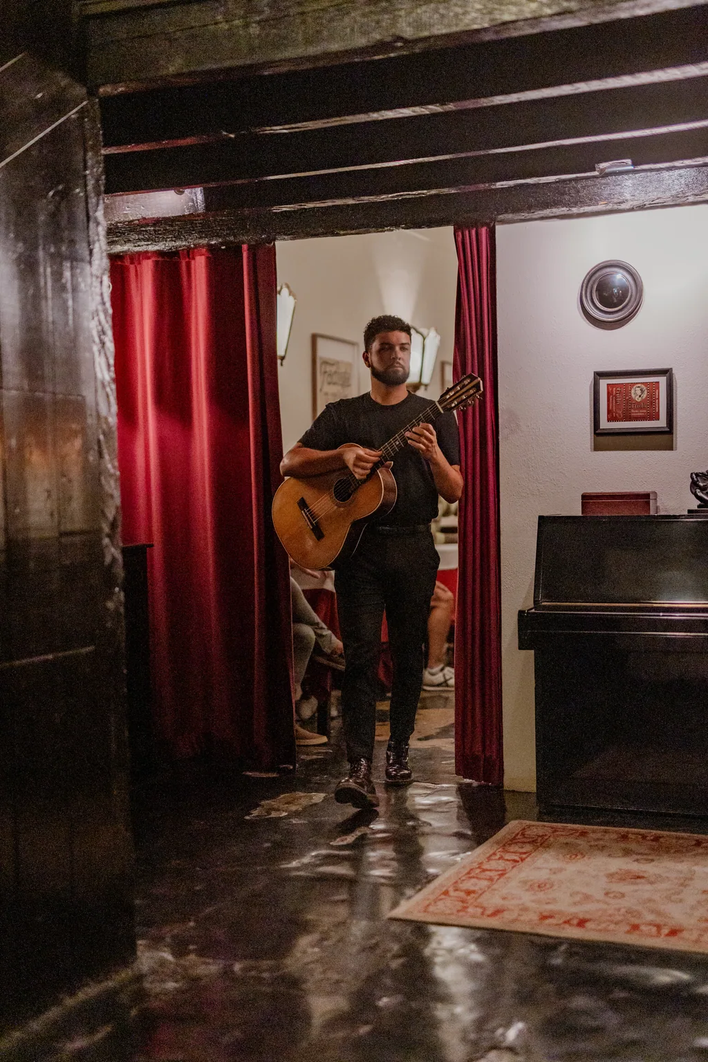Músico a atravessar a sala com a guitarra na mão, entre a cortina de veludo e o piano
